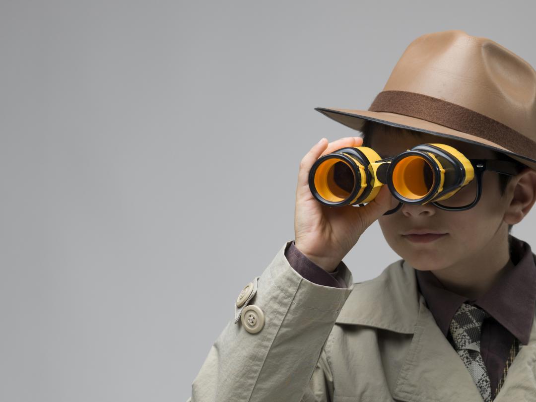 Een jongen in een trenchcoat kijkt door een verrekijker. Hij draagt een bril en een hoed.