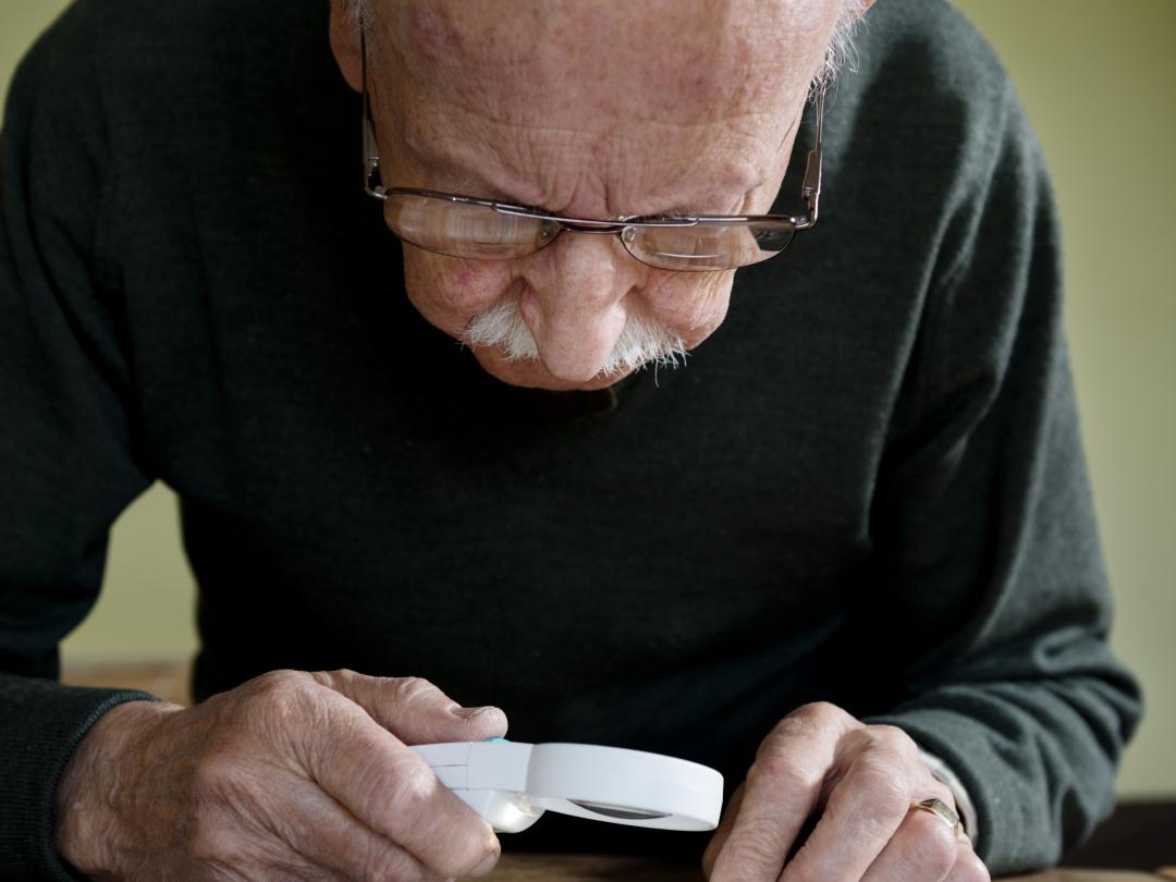 Een oude man staat voorover gebogen. Hij bestudeert iets met een vergrootglas.
