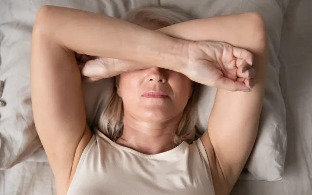 Een vrouw ligt op bed. Ze bedekt haar gezicht met haar armen kruislings over elkaar geslagen.