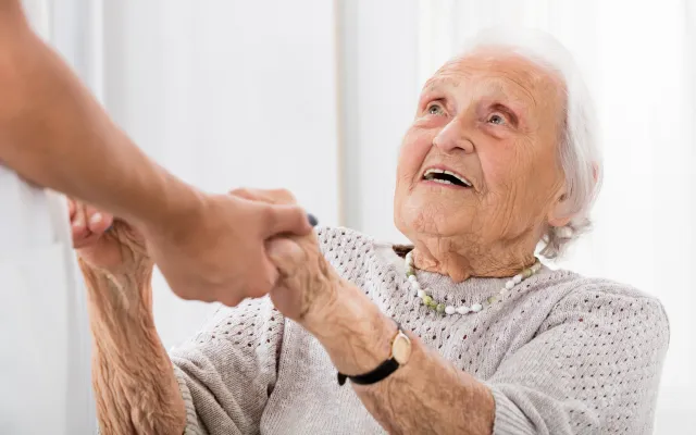 Een oude vrouw kijkt omhoog naar iemand die haar handen vasthoudt.