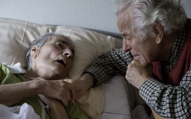 Twee oude mensen waarvan één op bed ligt. Ze houden elkaars hand vast.
