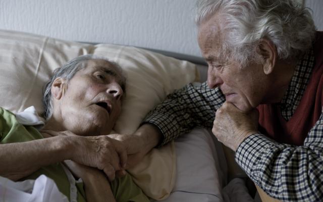 Twee oude mensen waarvan één op bed ligt. Ze houden elkaars hand vast.