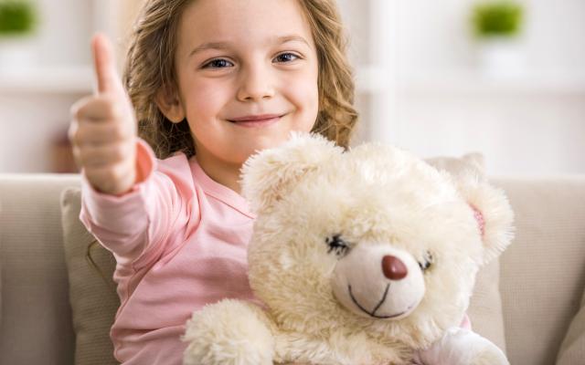 Een jong kind houdt een witte teddybeer vast en een duim omhoog.