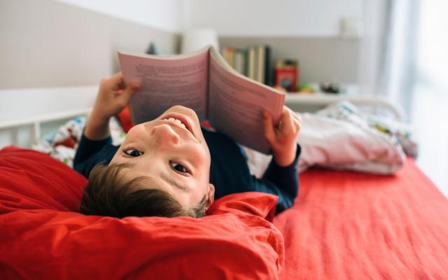 Een kind ligt op bed met een boek en kijkt vrolijk op naar de camera.