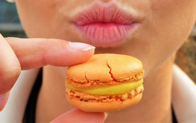Iemand houdt een oranje macaron voor haar getuite lippen.