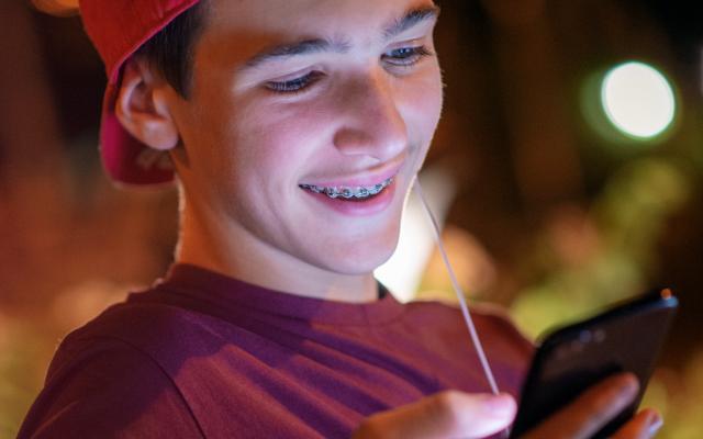 Een tiener met een beugel en een pet op kijkt lachend naar de telefoon in zijn hand.