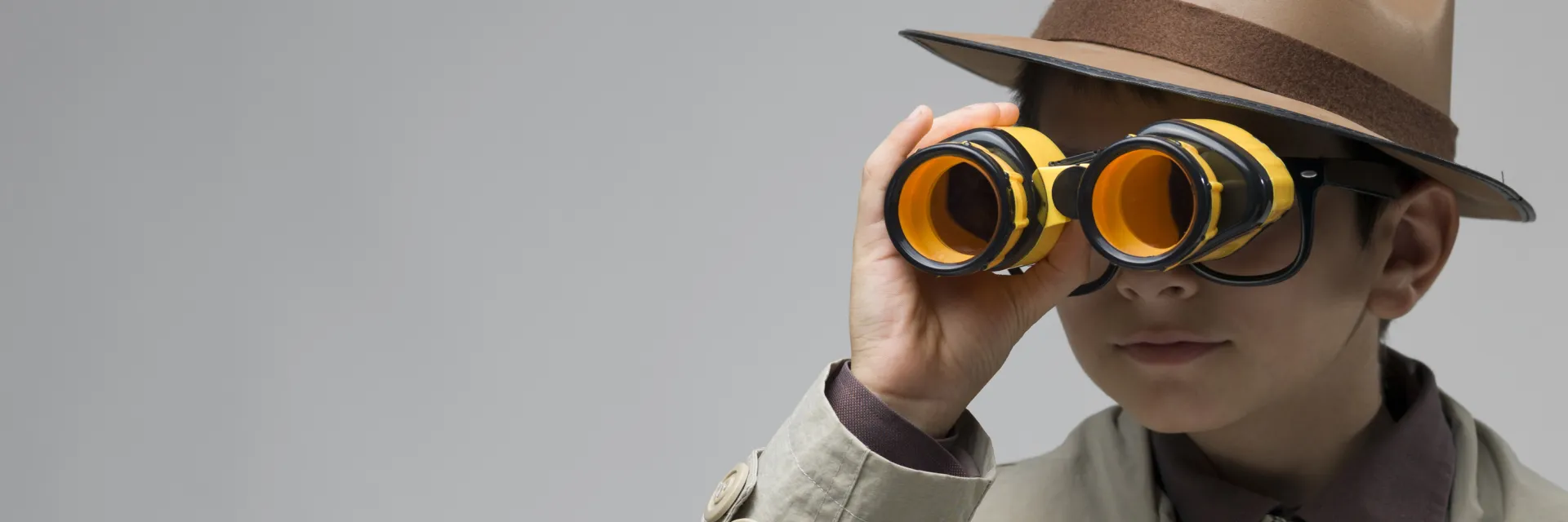 Een jongen in een trenchcoat kijkt door een verrekijker. Hij draagt een bril en een hoed.