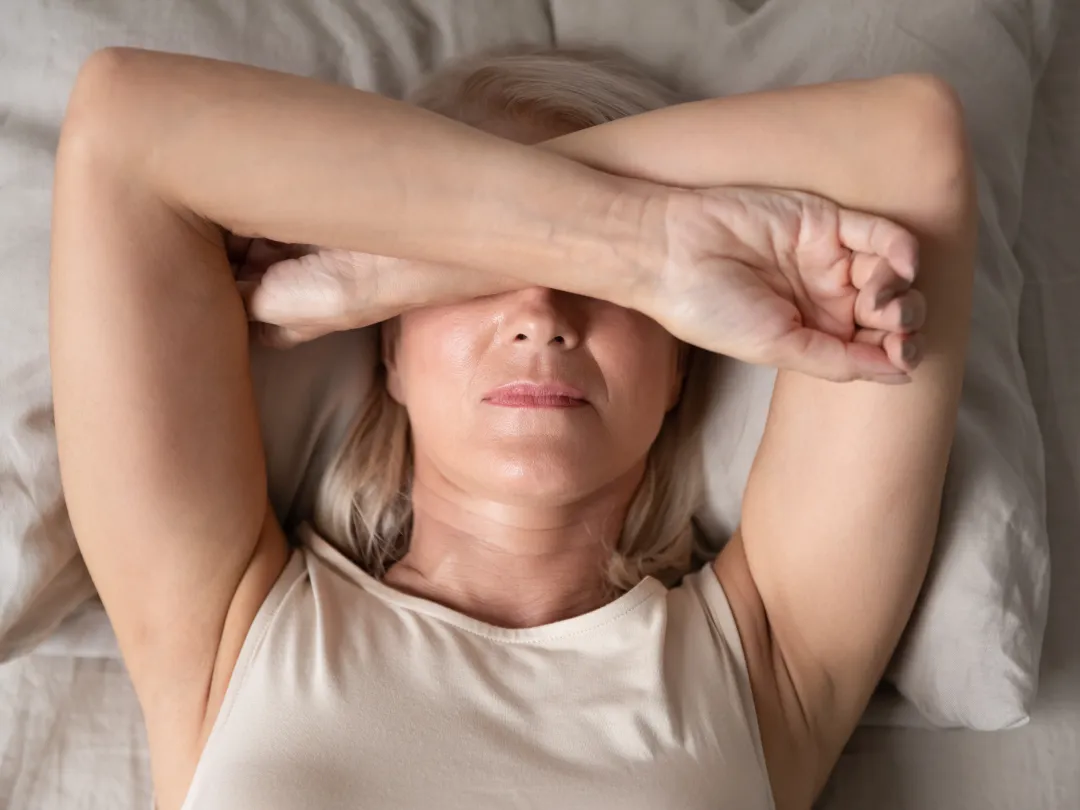 Een vrouw ligt op bed. Ze bedekt haar gezicht met haar armen kruislings over elkaar geslagen.