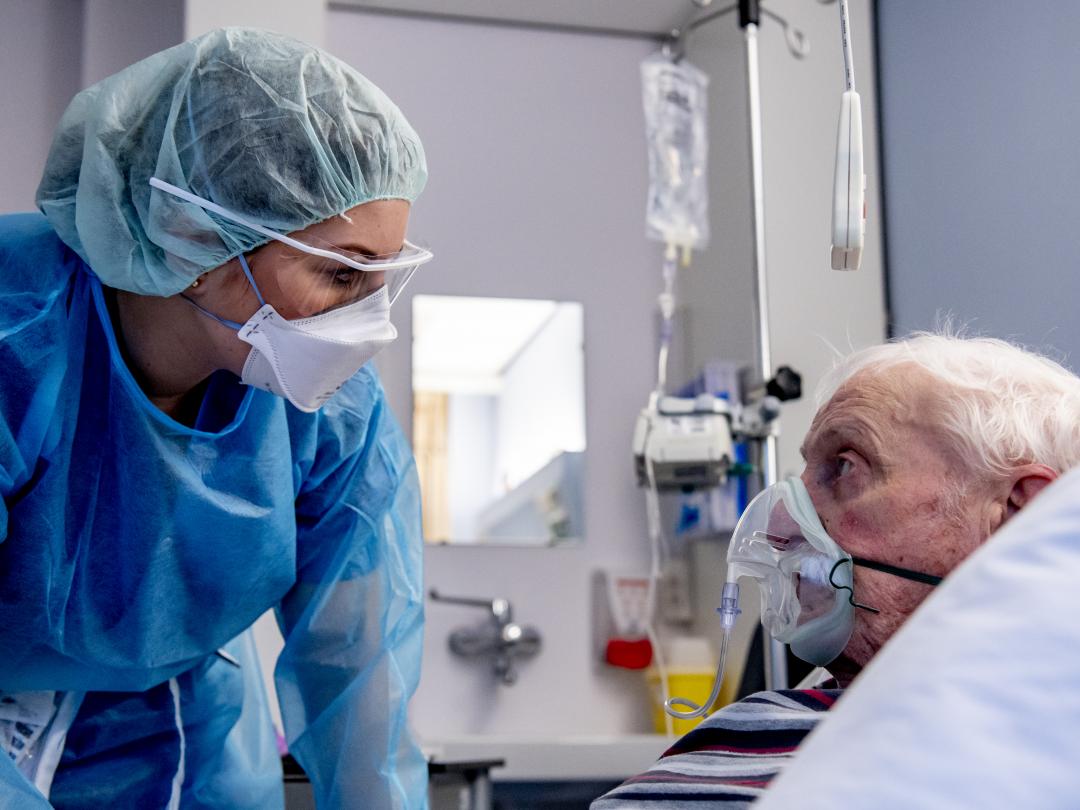Een arts of verpleegkundige draagt beschermende kleding en buigt zich over een oudere patiënt in een ziekenhuisbed.