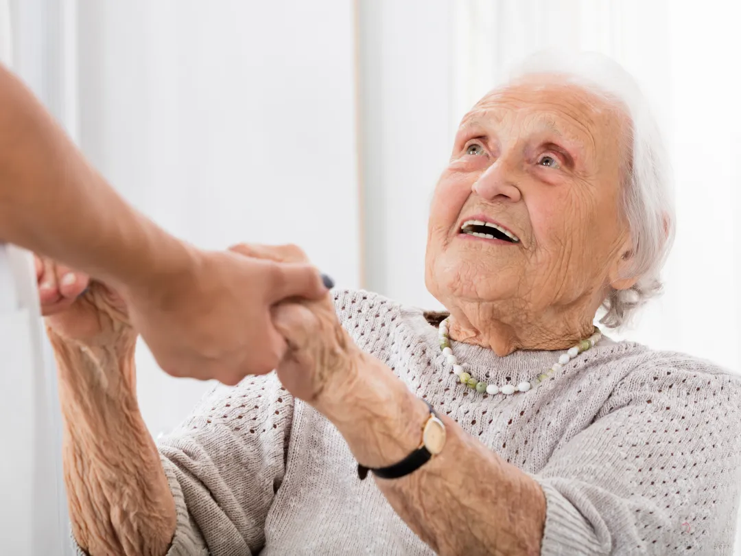 Een oude vrouw kijkt omhoog naar iemand die haar handen vasthoudt.
