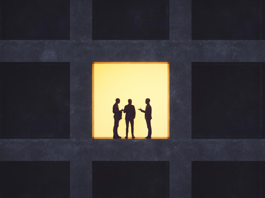 Drie silhouetten in gesprek in een verlicht raam van een verder donker gebouw.