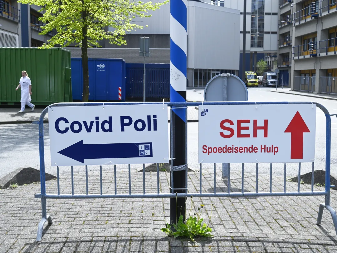 Een hek waar twee borden van het LUMC op bevestigd zijn. Op het linkerbord staat in het blauw 'Covid Poli' met een pijl naar links. OP het rechterbord staat in het rood 'SEH Spoedeisende Hulp' met een pijl naar boven.