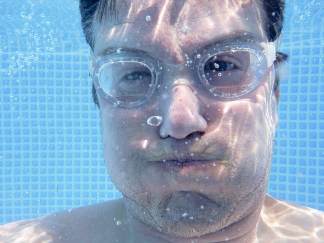 Gezicht van een man onderwater. Hij draagt een duikbril en heeft zijn wangen opgeblazen.