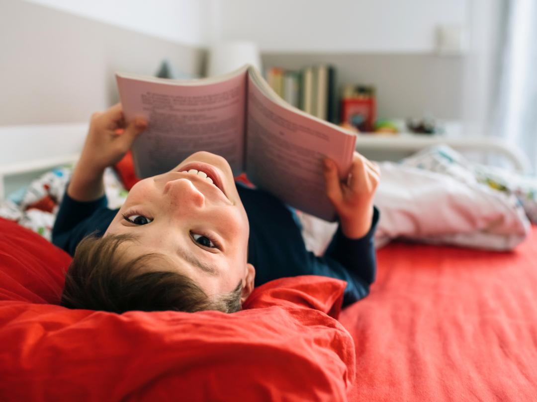 Een kind ligt op bed met een boek en kijkt vrolijk op naar de camera.