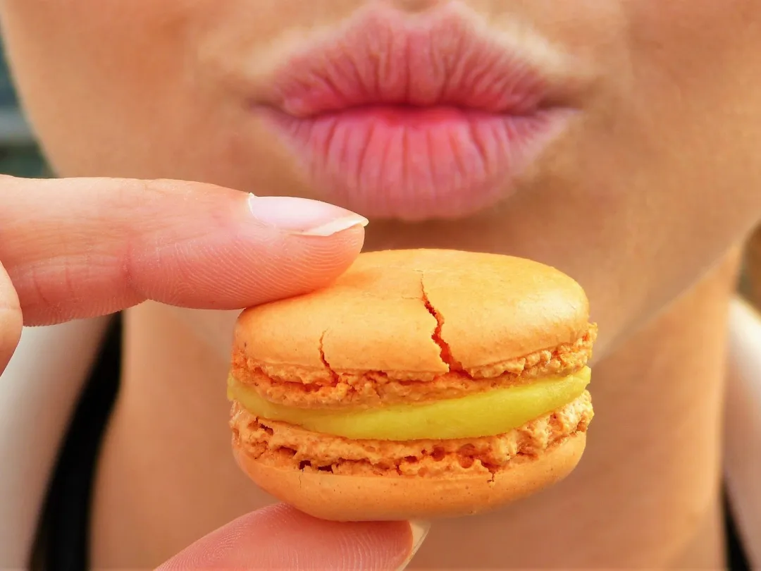 Iemand houdt een oranje macaron voor haar getuite lippen.