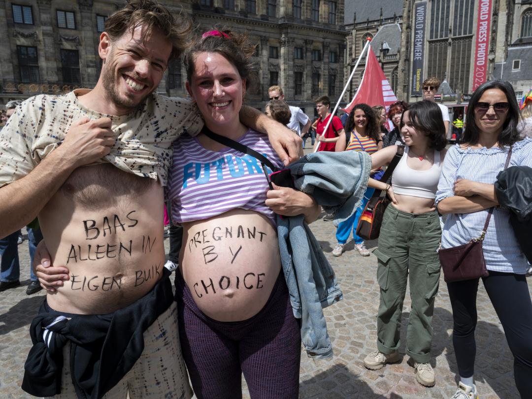 Mensen op de Dam in Amsterdam tijdens een demonstratie. Een stel met ontblote buiken lacht naar de camera. De man heeft op zijn buik de geschreven tekst 'baas alleen in eigen buik' staan, de vrouw draagt de tekst 'pregnant by choice'.