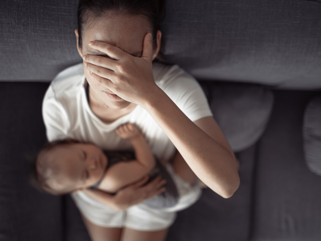 Een vermoeide vrouw die een slapende baby vasthoudt en met haar hand haar gezicht bedekt, wat een moment van uitputting of stress verbeeldt