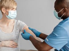 Een zwangere vrouw heeft haar mouw opgestroopt en krijgt een pleister op haar arm geplakt door een arts of verpleegkundige. Beiden dragen een mondkapje.