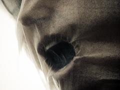 Er hangt een dunne stof over een gezicht heen. De mond van het gezicht is geopend en lijkt naar adem te happen.