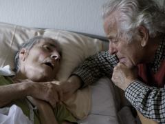 Twee oude mensen waarvan één op bed ligt. Ze houden elkaars hand vast.