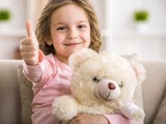 Een jong kind houdt een witte teddybeer vast en een duim omhoog.