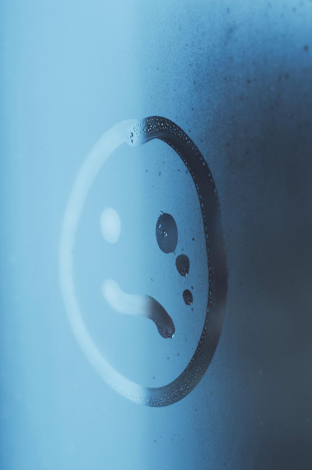 In condens op een raam is een smiley getekend met een half lachend en half huilend gezicht.
