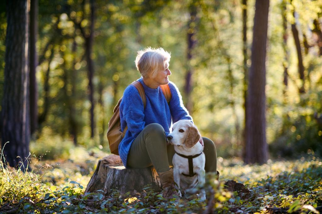 Vrouw met kort haar zit op een afgezaagde boomstronk met een rugzak om en haar linkerhand rustend op een labrador. 